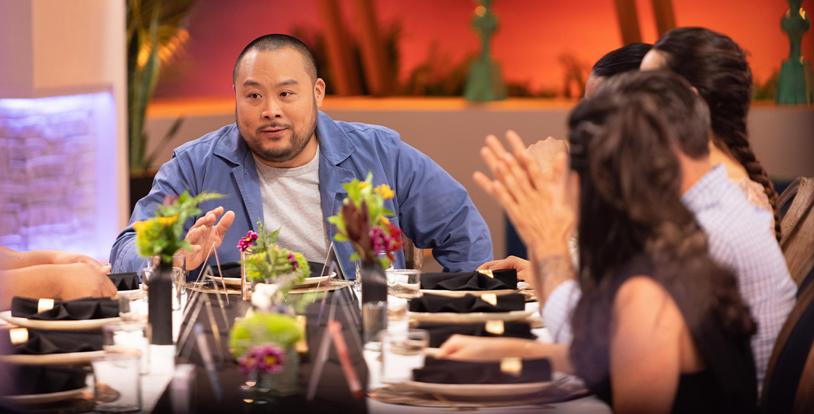 In an image from the new Hulu competition series Secret Chef, executive producer and chef David Chang is sitting at the head of a table, wearing a blue long-sleeved shirt with a white T-shirt underneath. Several of the show’s contestants are also sitting at the table, seen to his right—and there are plates, silverware, and flowers in vases in front of them; they are facing Chang as he speaks.