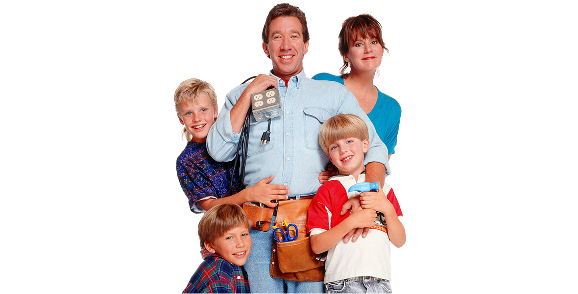 In a promotional image from ABC’s Home Improvement, now streaming on Disney+, Disney Legend Tim Allen is seen as Tim “The Tool Man” Taylor and is surrounded by his TV family: Patricia Richardson as Jill, Zachery Ty Bryan as Brad, Jonathan Taylor Thomas as Randy, and Taran Noah Smith as Mark. Allen is wearing a tool belt filled with tools and over his left shoulder is holding a square power strip. He is wearing a light denim button-up shirt and jeans. The rest of the family is wearing typical 1990s everyday clothing—flannel shirts, jeans, and the like. The whole family is seen against a white background.