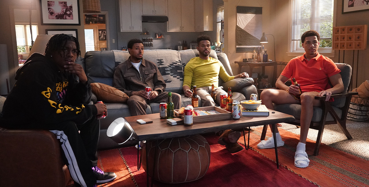 In an image from the Season 6 premiere of Freeform’s grown-ish, from left to right, Ceyair J. Wright as Zeke, Diggy Simmons as Doug, Marcus Scribner as Junior, and Trevor Jackson as Aaron are sitting around a living room, on chairs or a couch, watching an unseen TV. A coffee table is in front of them.