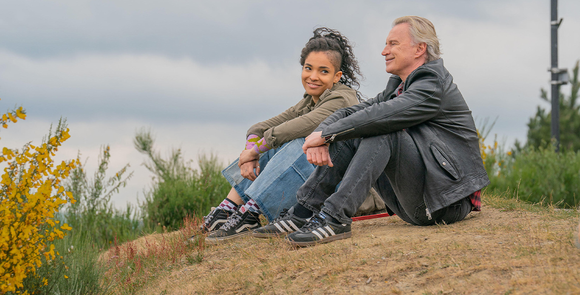 In an image from FX’s The Full Monty, Gaz (Robert Carlyle) and his teenage daughter Destiny (Talitha Wing) are sitting on a small hillside. It’s windy and overcast, and some flowers and tall grass surround them. Destiny is looking at her father and smiling; Gaz is looking forward. She wears an army green jacket and blue jeans; he wears a leather jacket and dark jeans.