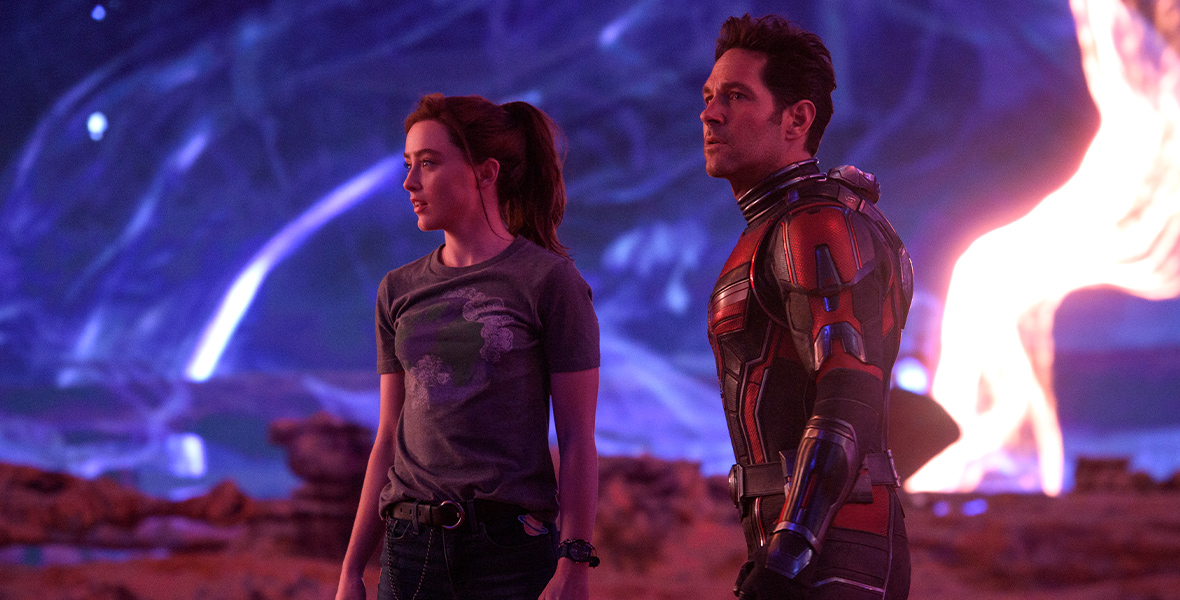 In an image from Marvel Studios’ Ant-Man and The Wasp: Quantumania, Cassie Lang (Kathryn Newton) and Scott Lang (Paul Rudd) are standing in the midst of Quantumania, looking at something to their left. There is a bright light to their right, and rock formations surround them below and in the distance.