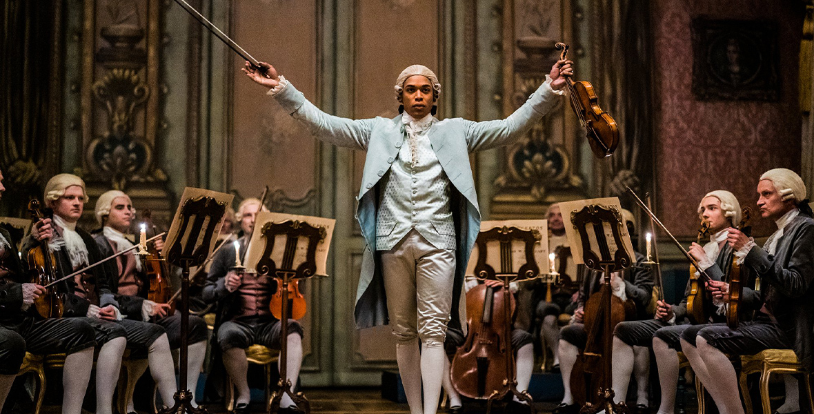 In an image from the Searchlight Pictures’ film Chevalier, Joseph Bologne (Kelvin Harrison Jr.) is standing amongst musicians, who are seated at either side of him. He is holding a violin and its bow aloft, as if he has just finished playing a piece. The musicians—some with violins, a few with cellos—all have music stands, some with candles attached, in front of them. All are wearing clothing specific to the French Revolution period.