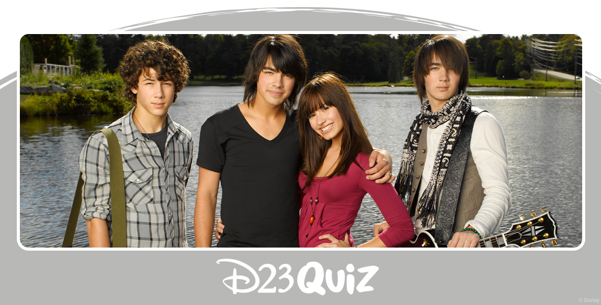 In an image from the Disney Channel Original Movie Camp Rock, from left to right, Nick Jonas, Joe Jonas, Demi Lovato, and Kevin Jonas are standing on a wooden dock at a lake. Nick and Kevin have guitars strung around their torsos. There are some clouds in the sky above them, but the sun is still shining through.