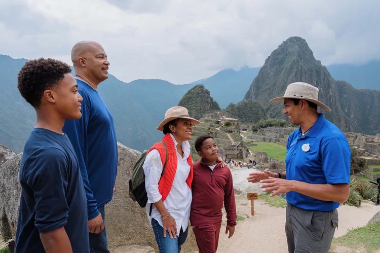 A tour guide talks with a family while standing in front of a mountain scape.