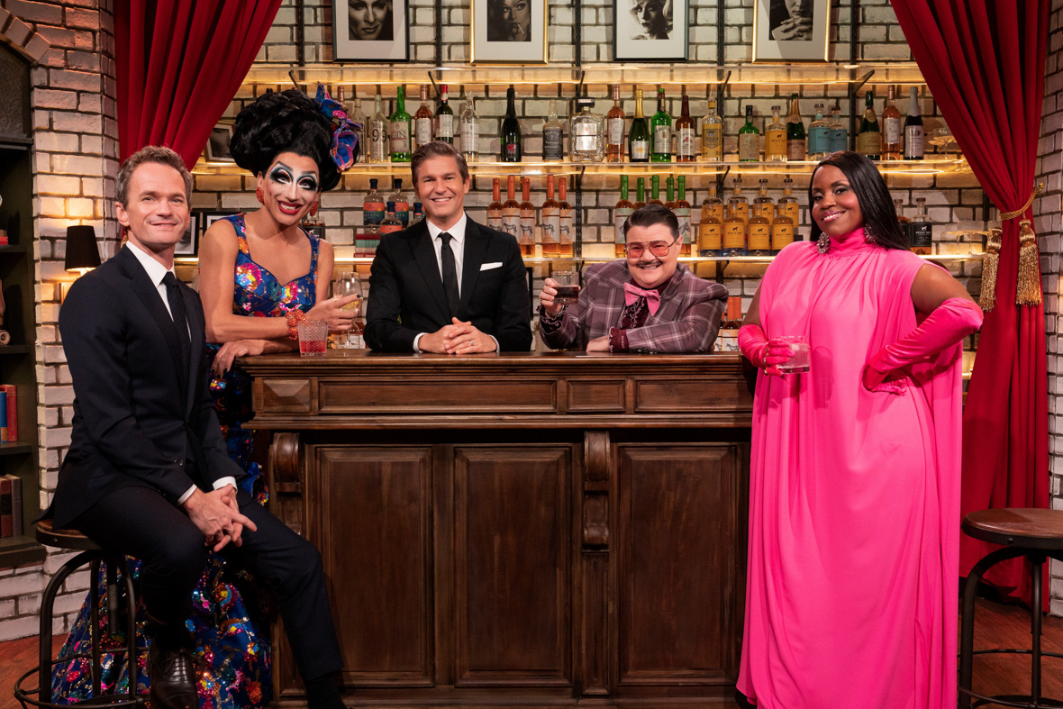 In this promo image for Hulu’s Drag Me to Dinner, from left to right, Neil Patrick Harris, Bianca Del Rio, David Burtka, Murray Hill, and Haneefah Wood are standing at a bar; behind them is a wall of liquor bottles. Harris and Burtka are dressed in tuxes; Hill is in a colorful suit. Rio and Wood are in colorful gowns. They are all smiling towards the camera.