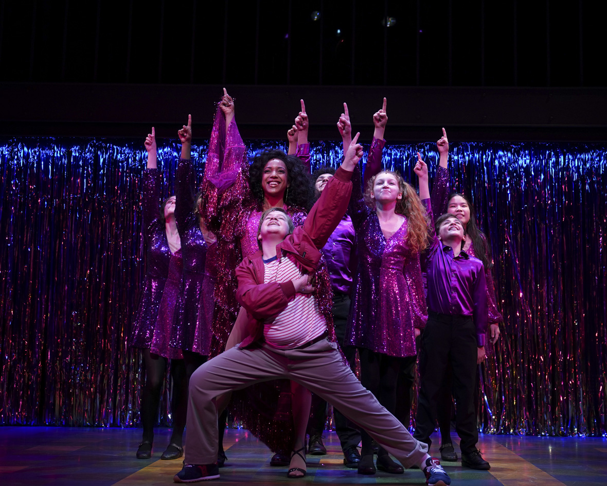 In this image from Trevor: The Musical, a filmed version of the Off-Broadway stage production, Holden William Hagelberger (as Trevor) and cast are in the middle of a musical number. Hagelberger is at center and is wearing a burgundy jacket, striped t-shirt, and gray pants. Behind him is someone portraying Diana Ross, wearing an appropriately glittery gown—and behind her is the rest of the cast, similarly garbed. Everyone has an arm raised in the air, pointing upwards; they’re all smiling and looking jubilant.