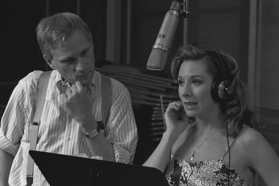 In a black and white still from the documentary Howard, Disney Legend Howard Ashman is seen standing next to Disney Legend Paige O’Hara at the microphone as she’s recording for Beauty and the Beast. Ashman is looking down at the pages on a music stand in front of O’Hara; O’Hara, wearing headphones, is looking up at the microphone. 