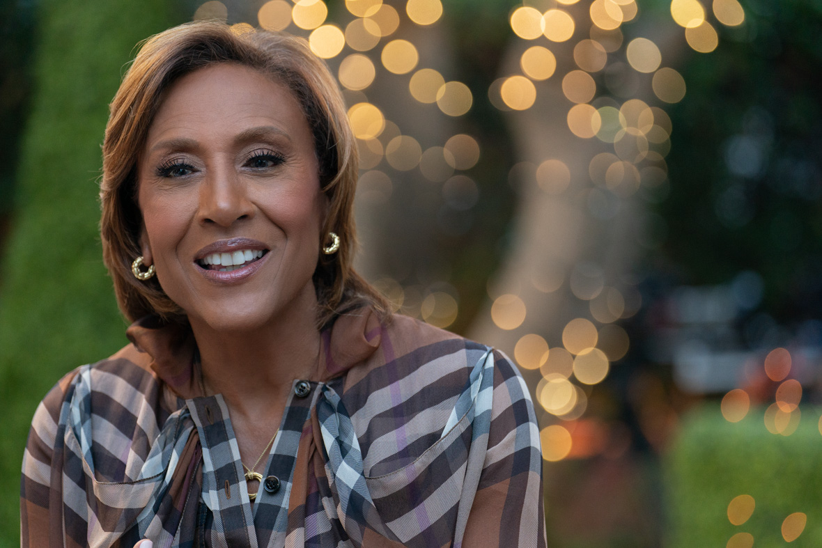An image of Disney Legend Robin Roberts from the Disney+ series Turning the Tables with Robin Roberts. She is wearing a black and white striped blouse, and blurry twinkly lights can be seen behind her.