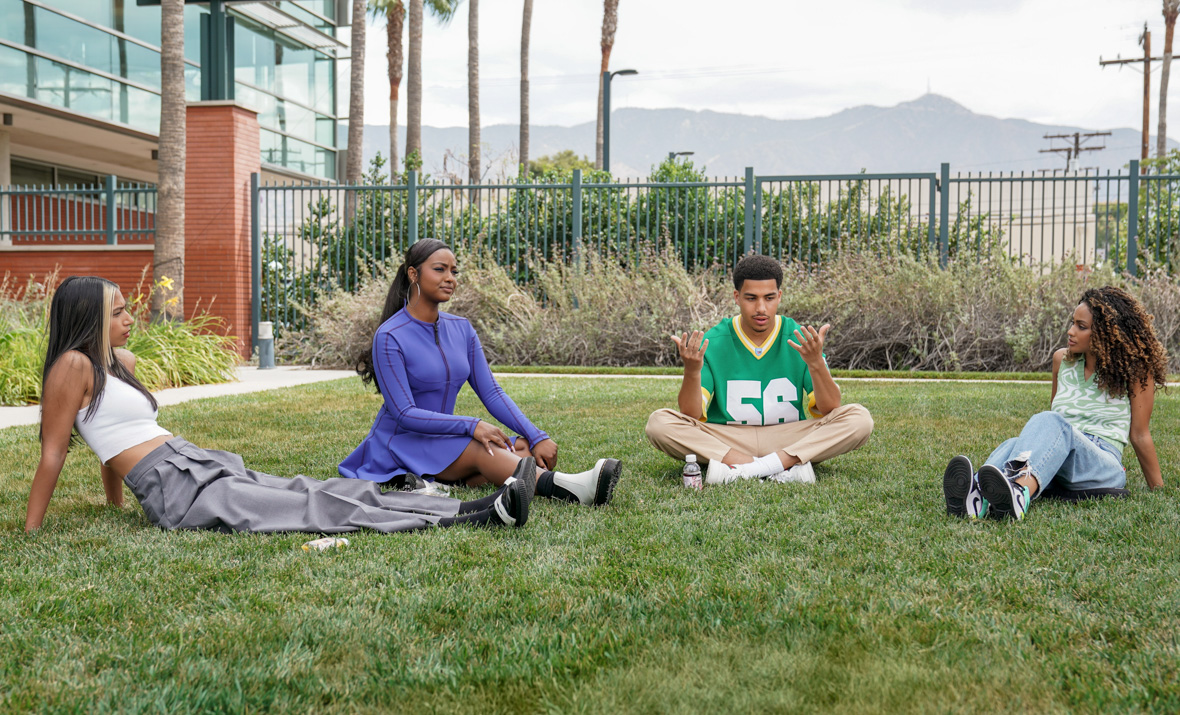 In an image from Freeform’s grown-ish, from left to right, Zaara (Tara Raani), Annika (Justine Skye), Junior (Marcus Scribner), and Kiela (Daniella Perkins) are sitting on the grass in front of a building at their college and chatting. They’re all wearing casual clothing.