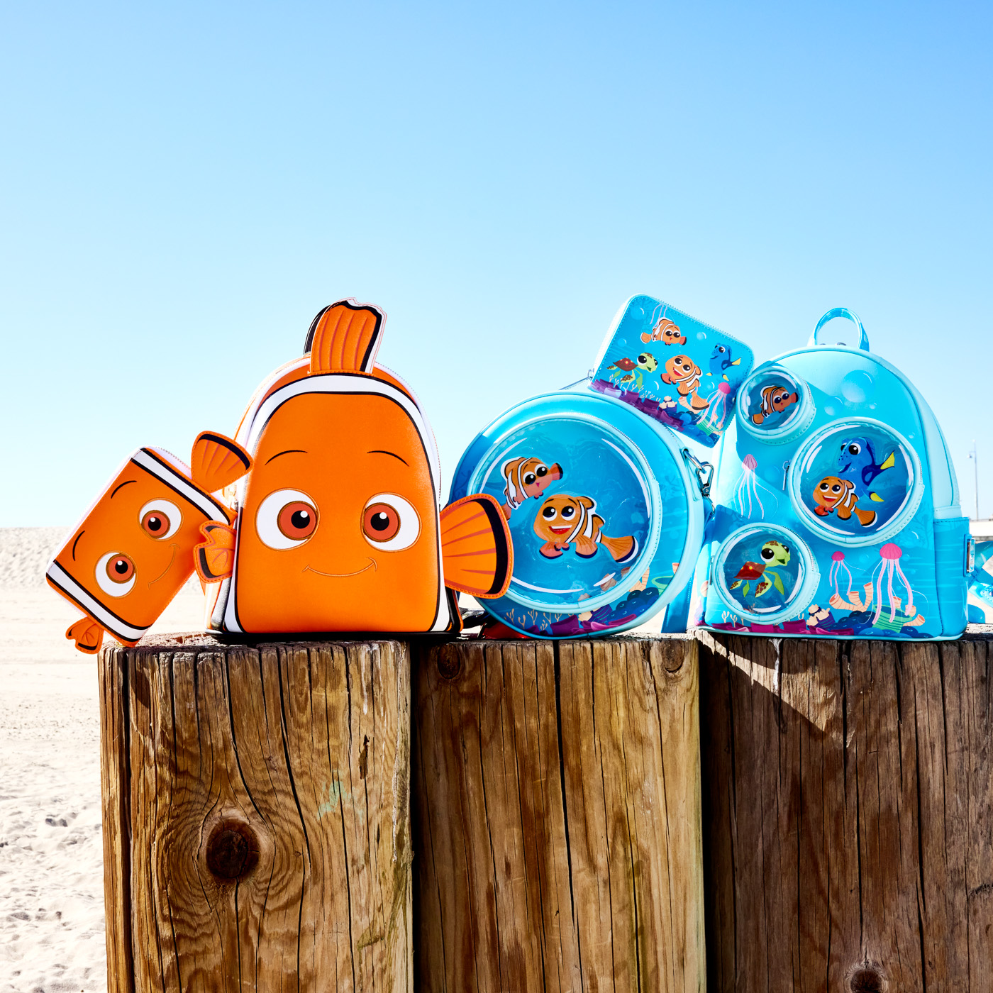 A bag and wallet designed to resemble Nemo, an orange clown fish. Next to it are two bags and a wallet, all featuring stylized art of Nemo, Marlin, Dory, and Crush swimming in the ocean. All the bags sit on top of some wooden poles at the beach.