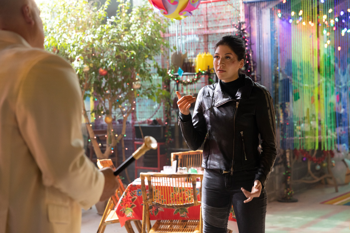 Maya Lopez, played by Alaqua Cox, uses American Sign Language (ASL) to communicate with Kingpin (Vincent D’Onofrio), in a scene from Marvel Studios’ Hawkeye. Maya is wearing a black leather jacket and skintight jeans, and her hair is pulled back. She has cuts and bruises on her face. Kingpin, whose back is turned to the camera, is holding a cane and wearing a white blazer. They are standing inside a colorful restaurant that is decorated for Christmas with garland, ornaments, and more.