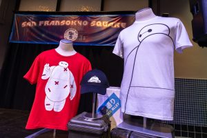 In this photo of a merchandise display, a small red shirt features Baymax with red bandages all over him, a larger white shirt sports a black outline of Baymax, and a black ball cap shows Baymax lying down with Mochi the cat lying on his stomach.