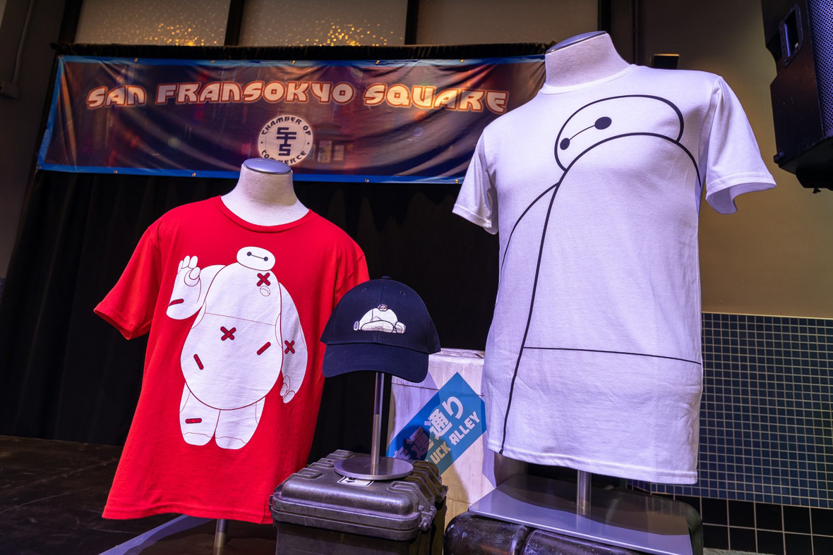 In this photo of a merchandise display, a small red shirt features Baymax with red bandages all over him, a larger white shirt sports a black outline of Baymax, and a black ball cap shows Baymax lying down with Mochi the cat lying on his stomach.