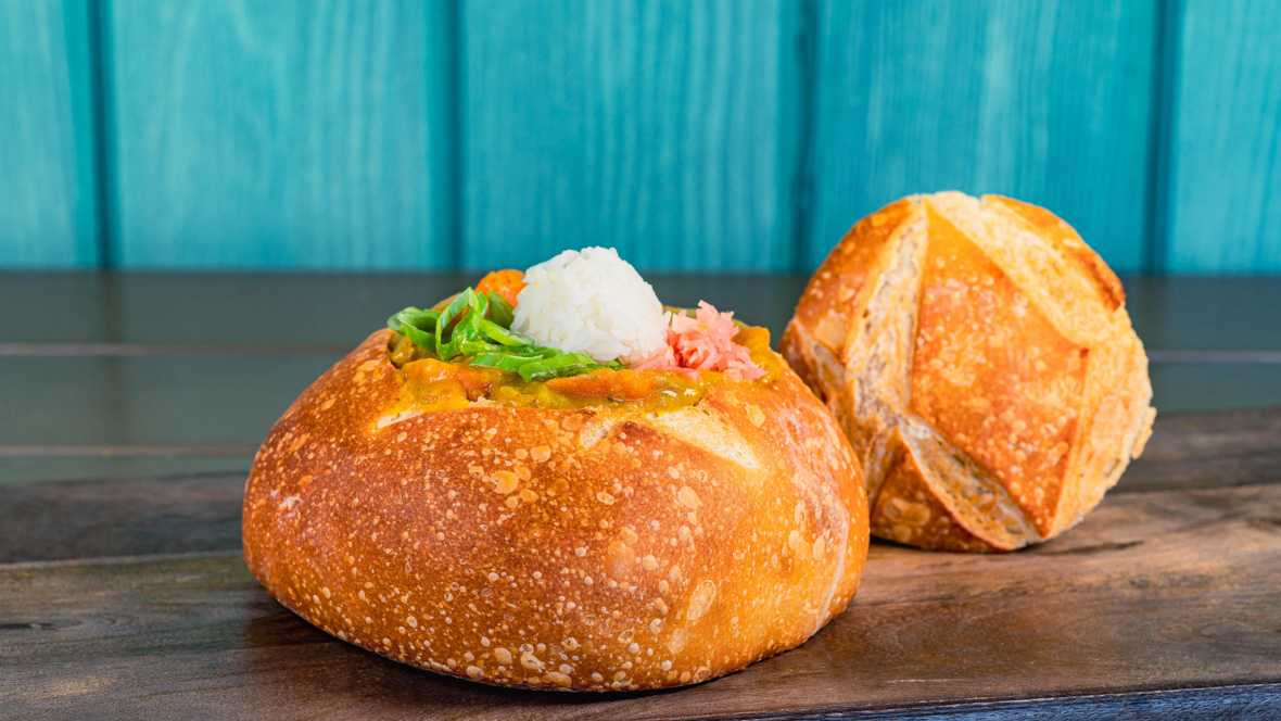 A bread bowl sits on a wooden table with a blue wall behind it. Inside is a curry with a rice ball on top, pickled ginger, and chopped green onion.