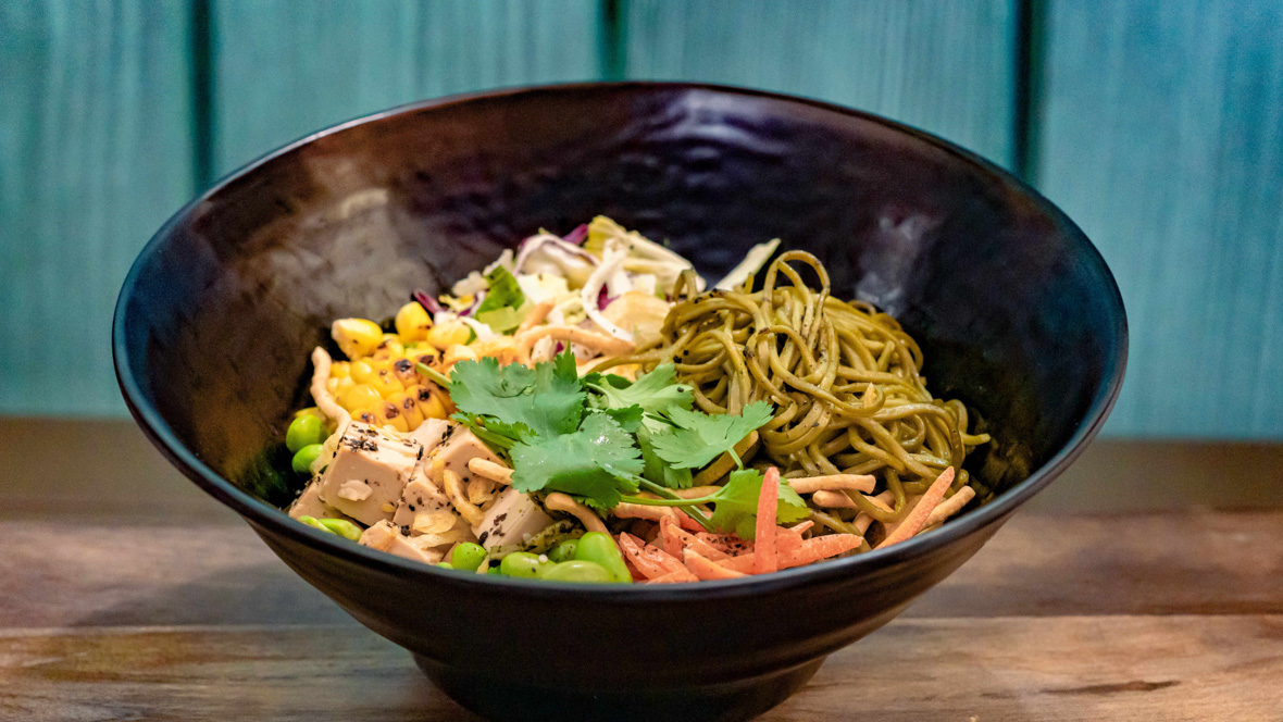 Soba noodles, squares of tofu, edamame, corn, slaw, carrots, crispy shallots, and cilantro all sit in a black bowl on a wooden table in front of a blue wall.