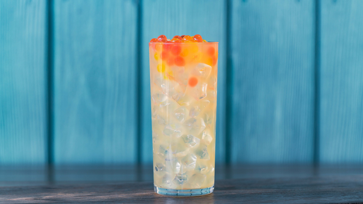 Lemonade is in a tall clear glass topped with ice and pink and orange popping boba on a wooden table in front of a blue wall.