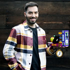 Marocs Bessa stands in front of a dark, wooden background, smiling as he holds up the LEGO Disney Villain set in his left hand.