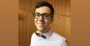 An image depicting LEGO designer Illia Gotlib who is wearing a dress shirt with thin blue stripes and a blue and red bow tie. He sports black-rimmed glassed and is smiling.