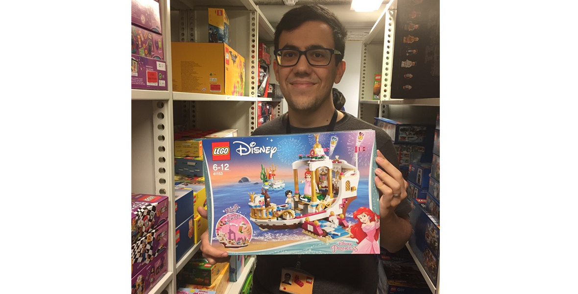LEGO Designer Illia Gotlib standing in a hallway with shelves of boxed LEGO sets. He is smiling and holding the box for a The Little Mermaid-themed LEGO set featuring Ariel and Prince Eric on a boat.