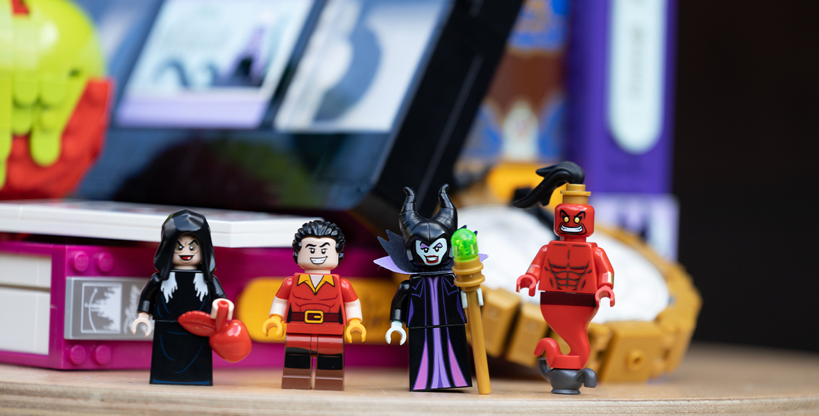 LEGO minifigures of the Evil Queen (Old Hag version) from Snow White, Gaston from Beauty and the Beast, Maleficent from Sleeping Beauty, and Jafar (Evil Genie version) stand in a row in front of the LEGO Disney Villains set.