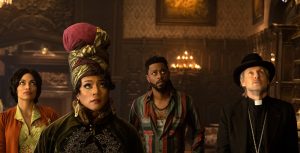 In an image from Disney’s Haunted Mansion, from left to right, Gabbie (Rosario Dawson), Harriet (Tiffany Haddish), Ben (LaKeith Stanfield), and Father Kent (Owen Wilson) are inside one of the rooms of the mansion and looking up at something offscreen, to the right. They have slightly worried looks on their faces. Harriet is wearing an elaborate headwrap, and Father Kent is wearing a black fedora and his usual priest collar.