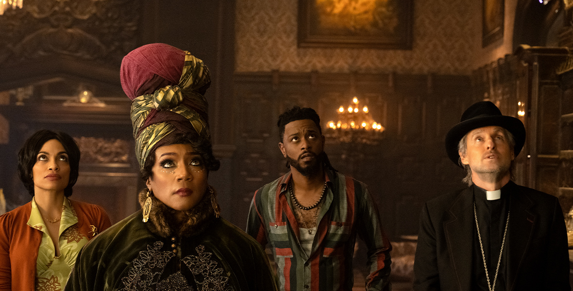 In an image from Disney’s Haunted Mansion, from left to right, Gabbie (Rosario Dawson), Harriet (Tiffany Haddish), Ben (LaKeith Stanfield), and Father Kent (Owen Wilson) are inside one of the rooms of the mansion and looking up at something offscreen, to the right. They have slightly worried looks on their faces. Harriet is wearing an elaborate headwrap, and Father Kent is wearing a black fedora and his usual priest collar.