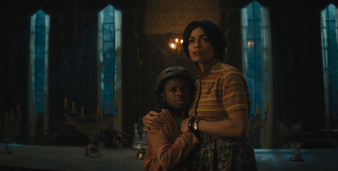 In an image from Disney’s Haunted Mansion, from left to right, Travis (Chase W. Dillon) is holding onto his mother Gabbie (Rosario Dawson) in a darkened room inside the mansion. They both have worried looks on their faces and are looking at something offscreen to the left. Travis is wearing a bicycle helmet.