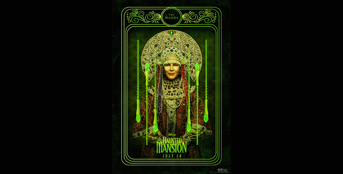 In a promotional image from Disney’s Haunted Mansion that looks like a tarot card for “The Wands,” Madame Leota (Jamie Lee Curtis) smiles at the camera wearing an elaborate headdress and jeweled high-neck dress, surrounded on either side by green glowing wands, all set against a black background. The logo for the film is at the bottom of the image, along with the date of the film’s release.