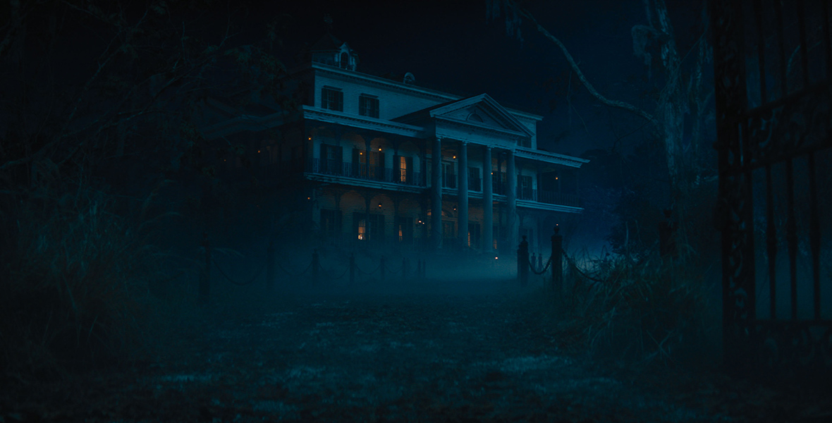 In an image from Disney’s Haunted Mansion, the mansion itself is seen at night, shrouded in fog. A driveway leads up to the front, denoted by short wooden columns with chains attached on either side. Four columns are seen on the front of the building, and a few lights are on inside.