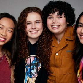 The leading cast from The Slumber Party, pose with their arms around each other. From left to right, Emmy Liu-Wang, who plays Paige, wears a dark red T-shirt with a light brown utility vest, Darby Camp, who plays Megan, is wearing a black sweatshirt with a blue, pink, and yellow skull on it, Alex Cooper Cohen, who plays Veronica, is wearing a burnt orange collared jumpsuit, and Valentina Herrera, who plays Anna Maria, is wearing a white shirt with a purple, pink, orange, and yellow logo on the front.