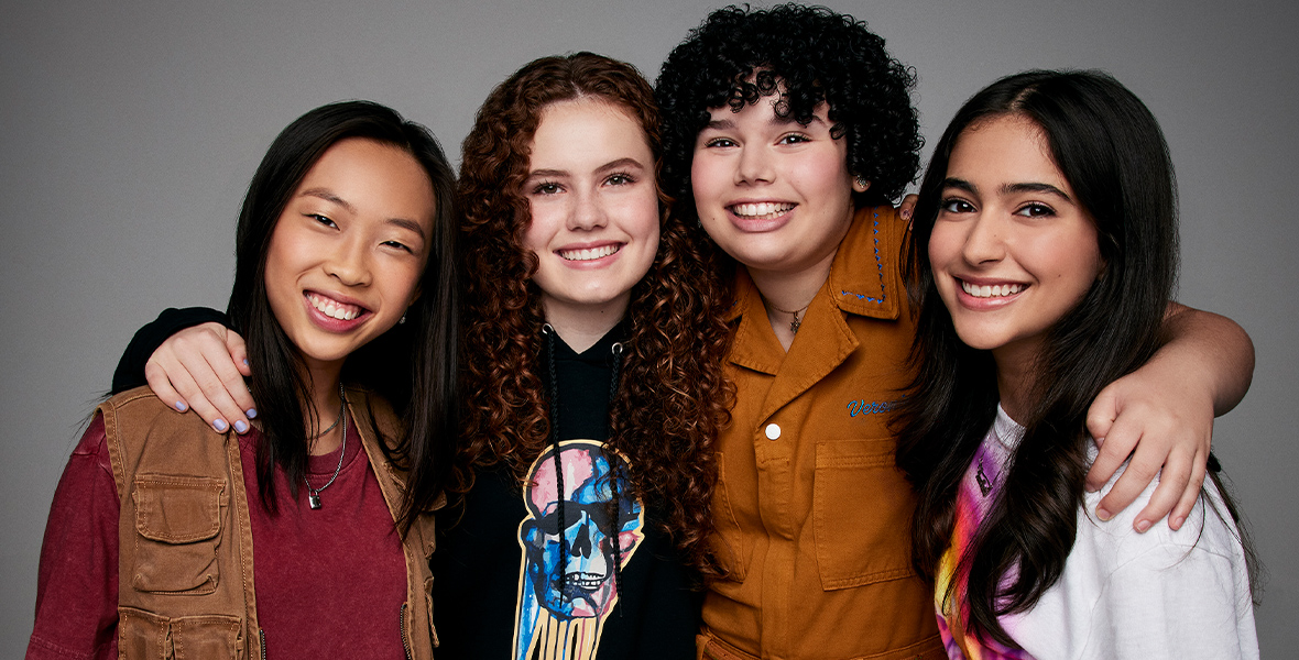 The leading cast from The Slumber Party, pose with their arms around each other. From left to right, Emmy Liu-Wang, who plays Paige, wears a dark red T-shirt with a light brown utility vest, Darby Camp, who plays Megan, is wearing a black sweatshirt with a blue, pink, and yellow skull on it, Alex Cooper Cohen, who plays Veronica, is wearing a burnt orange collared jumpsuit, and Valentina Herrera, who plays Anna Maria, is wearing a white shirt with a purple, pink, orange, and yellow logo on the front.