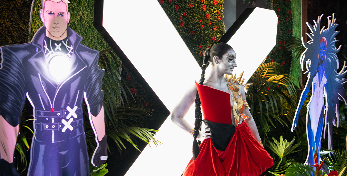 A guest posed in front of an illuminated X-Men logo on the green carpet at D23’s X-Men Hellfire Gala Event. They’re wearing an outfit inspired by Marvel Comics character Rasputin IV.
