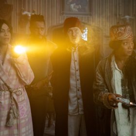 In an image from Disney’s Haunted Mansion, from left to right, Travis (Chase W. Dillon), Gabbie (Rosario Dawson), Ben (LaKeith Stanfield), Father Kent (Owen Wilson), and Harriet (Tiffany Haddish) are dressed in pajamas and huddled together looking for something in the mansion. Gabbie holds a flashlight that illuminates some sort of mist in front of the group. Everyone has concerned looks on their faces.