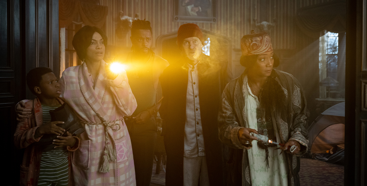 In an image from Disney’s Haunted Mansion, from left to right, Travis (Chase W. Dillon), Gabbie (Rosario Dawson), Ben (LaKeith Stanfield), Father Kent (Owen Wilson), and Harriet (Tiffany Haddish) are dressed in pajamas and huddled together looking for something in the mansion. Gabbie holds a flashlight that illuminates some sort of mist in front of the group. Everyone has concerned looks on their faces.