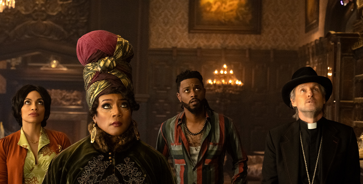 In an image from Disney’s Haunted Mansion, from left to right, Gabbie (Rosario Dawson), Harriet (Tiffany Haddish), Ben (LaKeith Stanfield), and Father Kent (Owen Wilson) are standing in one of the mansion’s large rooms and are looking up toward the right at the ceiling. They have concerned looks on their faces. Harriet is wearing an elaborate headwrap, and Father Kent is wearing a black fedora and other ecumenical garb.