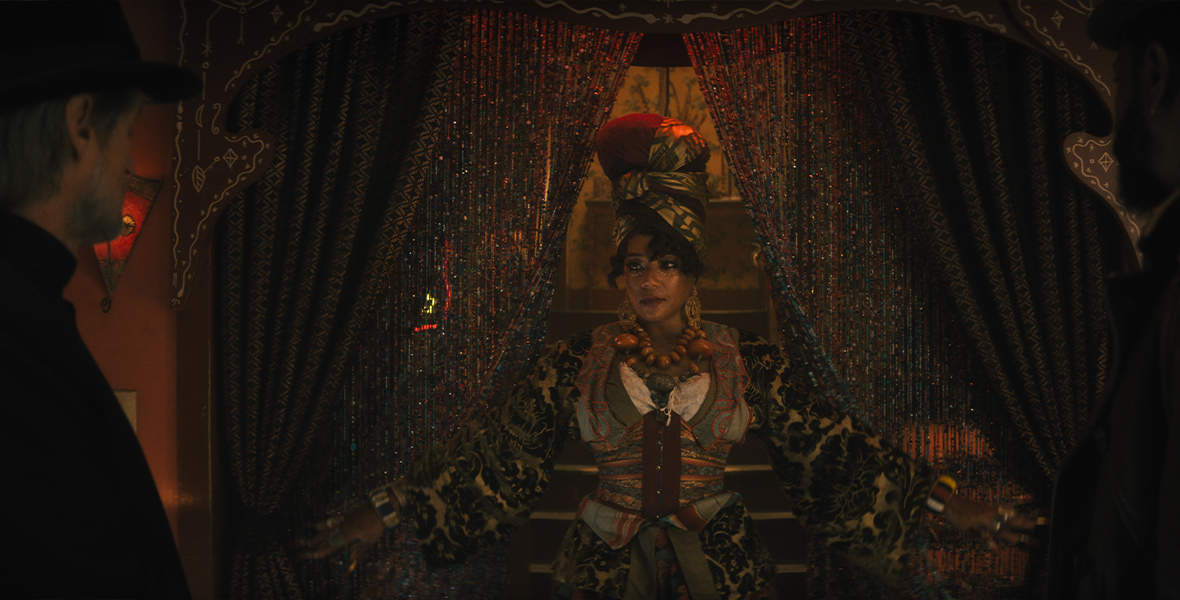 In an image from Disney’s Haunted Mansion, Harriet (Tiffany Haddish) is coming through a beaded curtain, looking at Father Kent (Owen Wilson), who’s facing her—with his back towards the camera—at the left of the image. She is wearing an elaborate headwrap, several pieces of jewelry, and a patterned dress with a cinched vest over it. The room is dimly lit.