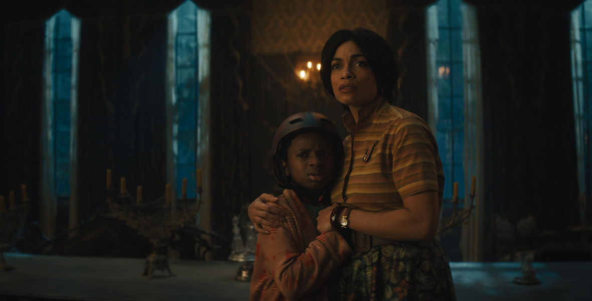 In an image from Disney’s Haunted Mansion, Travis (Chase W. Dillon) and Gabbie (Rosario Dawson) are huddled together, looking at something off-screen to the left. They have concerned looks on their faces. Travis is wearing a bike helmet and a robe; Gabbie is wearing an orange and yellow striped top and a flowered skirt. They appear to be in the mansion’s dining room, which is dimly lit; a long table is behind them, with some candelabras placed on top. Long thin windows can be seen in the background.