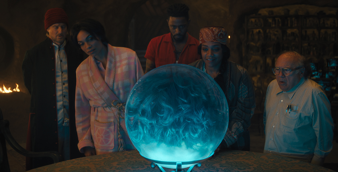 In an image from Disney’s Haunted Mansion, from left to right, Father Kent (Owen Wilson), Gabbie (Rosario Dawson), Ben (LaKeith Stanfield), Harriet (Tiffany Haddish), and Professor Davis (Danny DeVito) are standing behind a table, on which is displayed a large crystal ball. Inside the ball, we can see the back of someone’s ghostly head; while we can’t see her face, we know it is Madame Leota. Father Kent, Gabbie, Ben, and Harriet are wearing pajamas; Prof. Davis is wearing a button-up shirt with pens in the front pocket. Harriet is smiling. To the left of the group is some sort of fire pit or fireplace. 