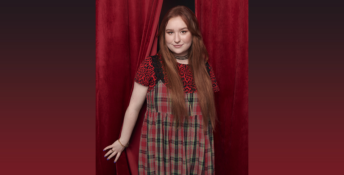 Julia Lester, who plays Ashlyn in High School Musical: The Musical: The Series, emerges from behind red curtains. She’s wearing a red, green, and tan plaid dress over a red-and-black short-sleeve top and has a triple-strand choker necklace around her neck.