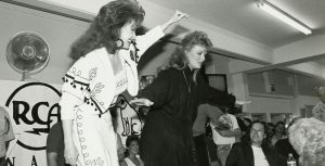 In a black-and-white image from Hulu&rsquo;s CMA Fest: 50 Years of Fan Fair, Naomi and Wynonna Judd stand on chairs behind a table during an autograph signing at a past CMA Fest. Fans gather around the table. Naomi is waving with one arm, while Wynonna has both of her arms outstretched. Naomi is wearing a light-colored outfit, while Wynonna&rsquo;s is darker; both have long, late &rsquo;80s/early &rsquo;90s-looking hair. A sign with the RCA logo can be seen behind them.