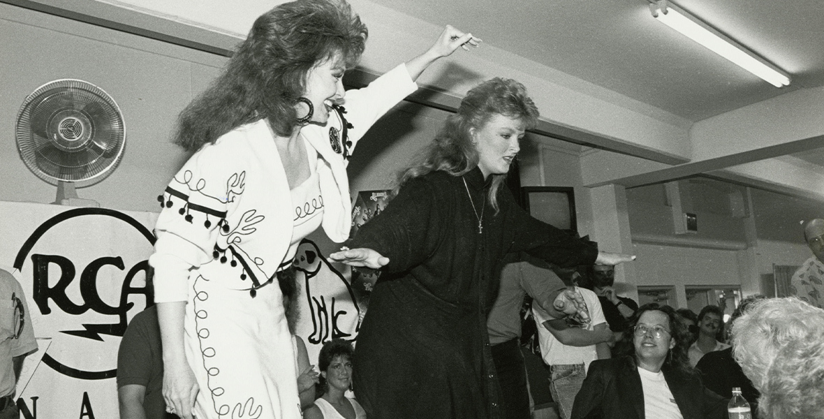 In a black-and-white image from Hulu’s CMA Fest: 50 Years of Fan Fair, Naomi and Wynonna Judd stand on chairs behind a table during an autograph signing at a past CMA Fest. Fans gather around the table. Naomi is waving with one arm, while Wynonna has both of her arms outstretched. Naomi is wearing a light-colored outfit, while Wynonna’s is darker; both have long, late ’80s/early ’90s-looking hair. A sign with the RCA logo can be seen behind them.