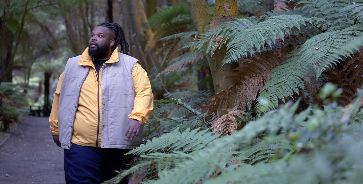 In an image from Never Say Never with Jeff Jenkins, Jenkins hikes in a thickly wooded area near the Lake Tarawera shore in Rotorua, New Zealand. He wears a light purple vest and yellow long-sleeved shirt and is surrounded by beautiful greenery. He looks around in awe.