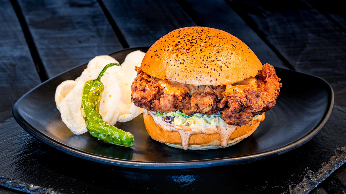 Sitting on a black plate is a fried chicken sandwich with a slaw and mayonnaise, a side of chips, and a grilled pepper. The black plate sits on a wooden table.