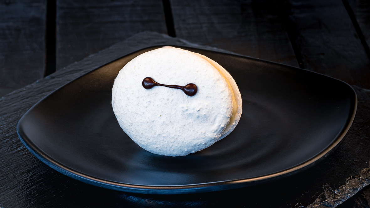 A large white macaron with a design representative of Baymax’s eyes sits on a black plate on a wooden table.