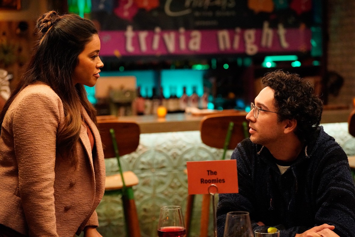 Nell, played by Gina Rodriquez stands to the left of a small table in profile mid-conversation. Her long brown hair is half-pulled up in a bun. She wears a peach-colored blazer over an orange blouse. A glass of red wine is on her side of the table. Her roommate Edward, played by Rick Glassman, sits at a table to her right and is looking at her. He is wearing wire-rim glasses and a woven sweatshirt. A wine glass and another drink glass with lime slice are visible near Edward. A placard on the table reads “The Roomies,” and is stationed in front of a long bar where a bright pink banner with white lettering announces: “trivia night.”