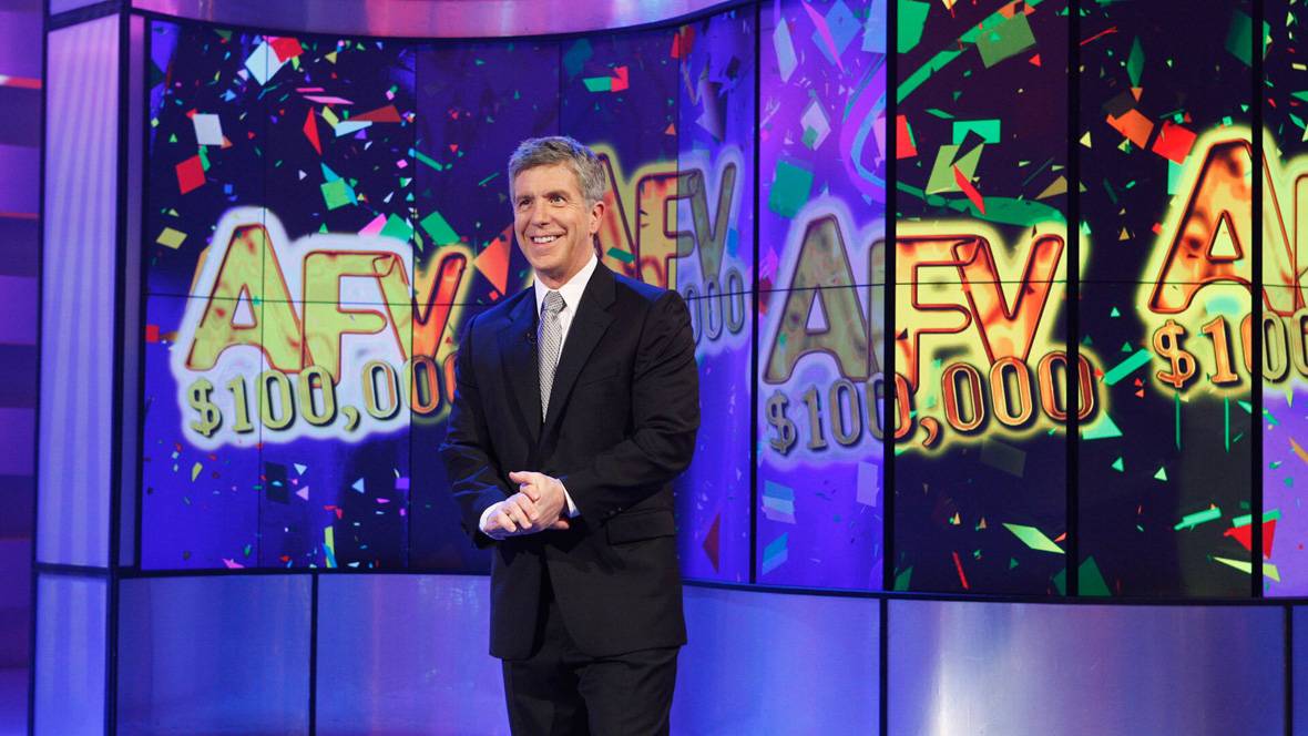 In an image from a past episode of America’s Funniest Home Videos, host Tom Bergeron, wearing a dark suit, stands in front of a purple-hued bank of video screens displaying the “AFHV” logo and “$100,000” multiple times, amongst digital confetti. Bergeron is smiling and looking off camera to the left.