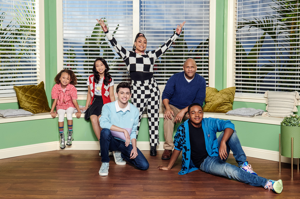 In a promo image for Disney Branded Television’s Raven’s Home are Raven-Symoné as Raven Baxter, Issac Ryan Brown as Booker Baxter-Carter, Rondell Sheridan as Victor Baxter, Mykal-Michelle Harris as Alice, Felix Avitia as Neil, and Emmy Liu-Wang as Ivy. They’re standing or sitting in a living room-style set with a large bay window; Raven-Symoné is wearing a black and white checkered crop top and pants and is holding her arms aloft, and everyone else is in casual clothing. They are all smiling at the camera.
