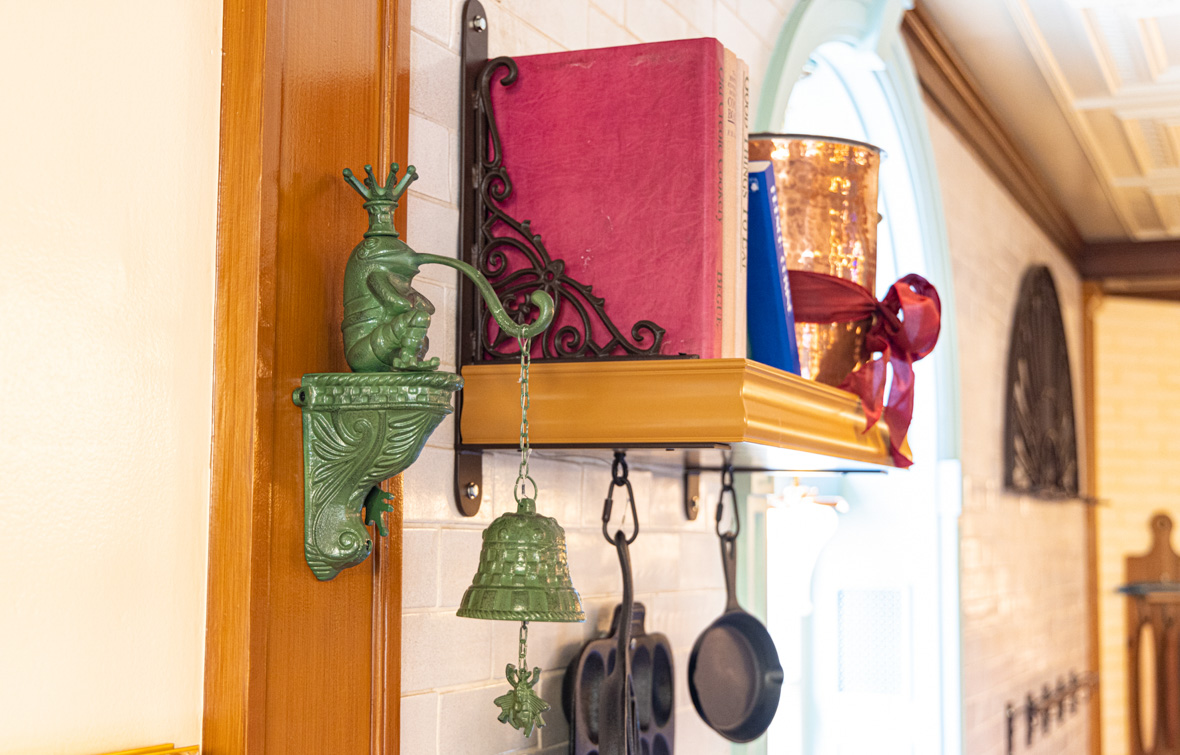 A small shelf hanging on the wall at Tiana’s Palace contains cookbooks on top and pots hanging from hooks below. A green porceline bell hangs to one side, in the shape of a frog prince.