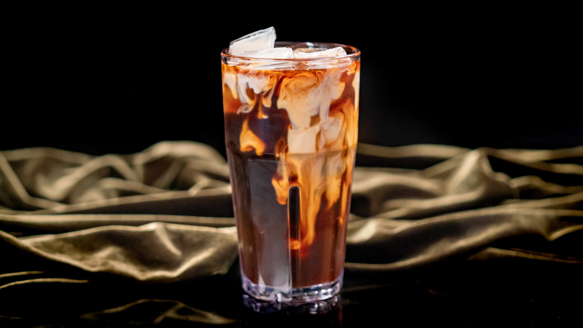 A chilled glass holds a serving of the Joffrey’s Coffee Chicory Cold Brew served at Tiana’s Palace. The glass shows swirls of cream and coffee colors and sits on a drak green cloth.