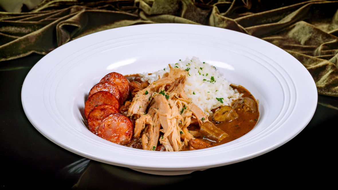 A bowl holds a serving of House Gumbo, as served at Tiana’s Palace. The dish features sliced sausage, shredded chicken, white rice, and vegetables.